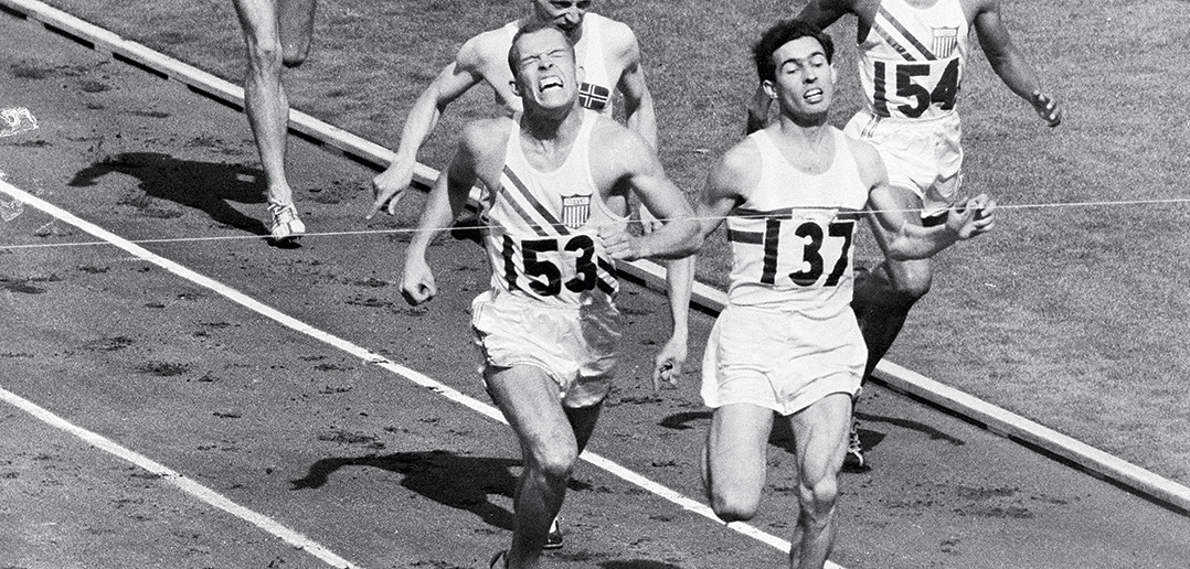 Sprinters on a track race toward the tape at the finish line, with one in expression of agony just edging out the others.