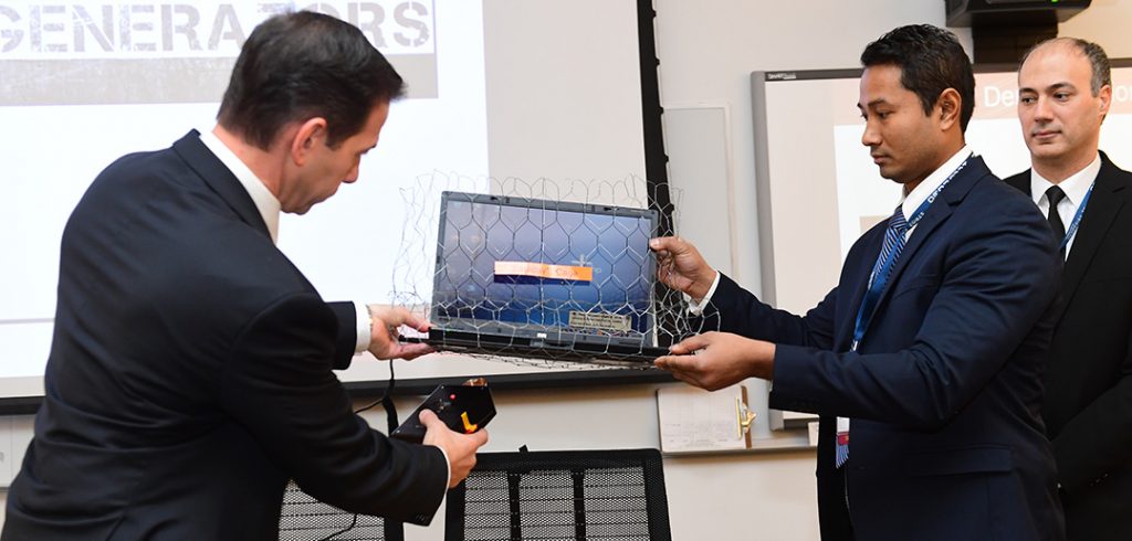 Damianos Pinou activates an electromagnetic pulse generator under a laptop wrapped in a metal mesh “Faraday cage” held by Rien Chy, as Thaier Hayajneh looks on.