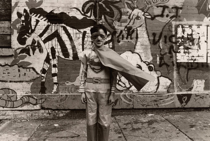 A young boy in wears a Superman costume and stands in front of a graffiti-tagged wall