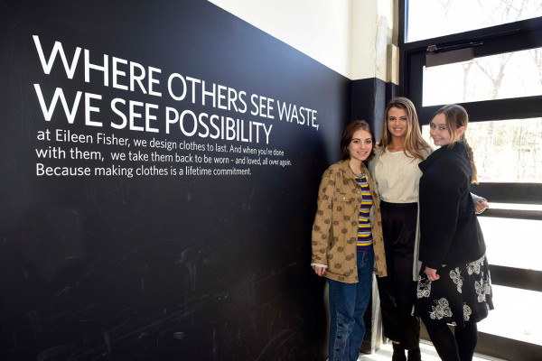 Three female students affiliated with the Social Innovation Collaboratory stand next to a black wall at the Eileen Fisher Company
