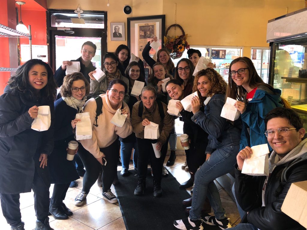 Gambito and her students stand together and smile in a pastry shop, holding white paper bags up to the camera. 