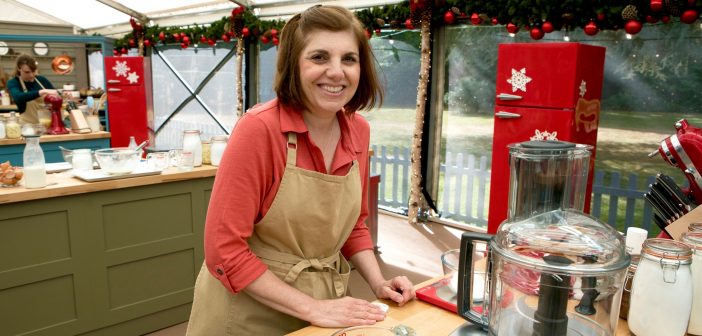 Tina Zaccardi, wearing a beige apron and a dark pink top, smiles for the camera in a kitchen.