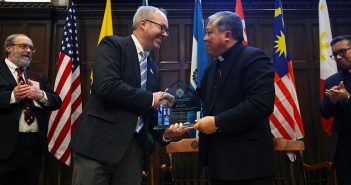 John Briggs shake hands with Archbishop Bernardito Auza on stage in the Keating First floor auditorium, with flags behind them.