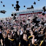 Graduates wearing black academic robes throw their hats in the air on the football field
