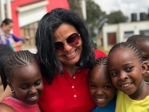 A Hispanic woman wearing a red shirt and sunglasses smiles and holds three young Kenyan children