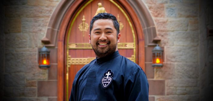 A man wearing a black priest robe smiles in front of a set of church doors