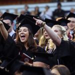 Students wearing black graduation gowns cheer and point their fingers