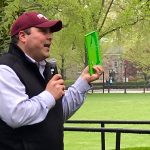 A man wearing a maroon Fordham baseball cap holds the book "The Giving Tree" in front of a backdrop of trees.