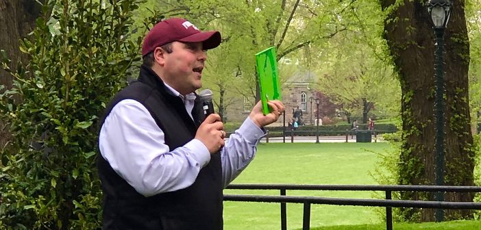 A man wearing a maroon Fordham baseball cap holds the book "The Giving Tree" in front of a backdrop of trees.