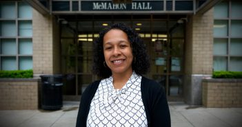 A woman wearing a blouse and a dark cardigan smiles in front of McMahon Hall.