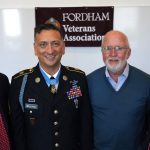 Four men smile and stand together in front of a flag that says "Fordham Veterans Association."