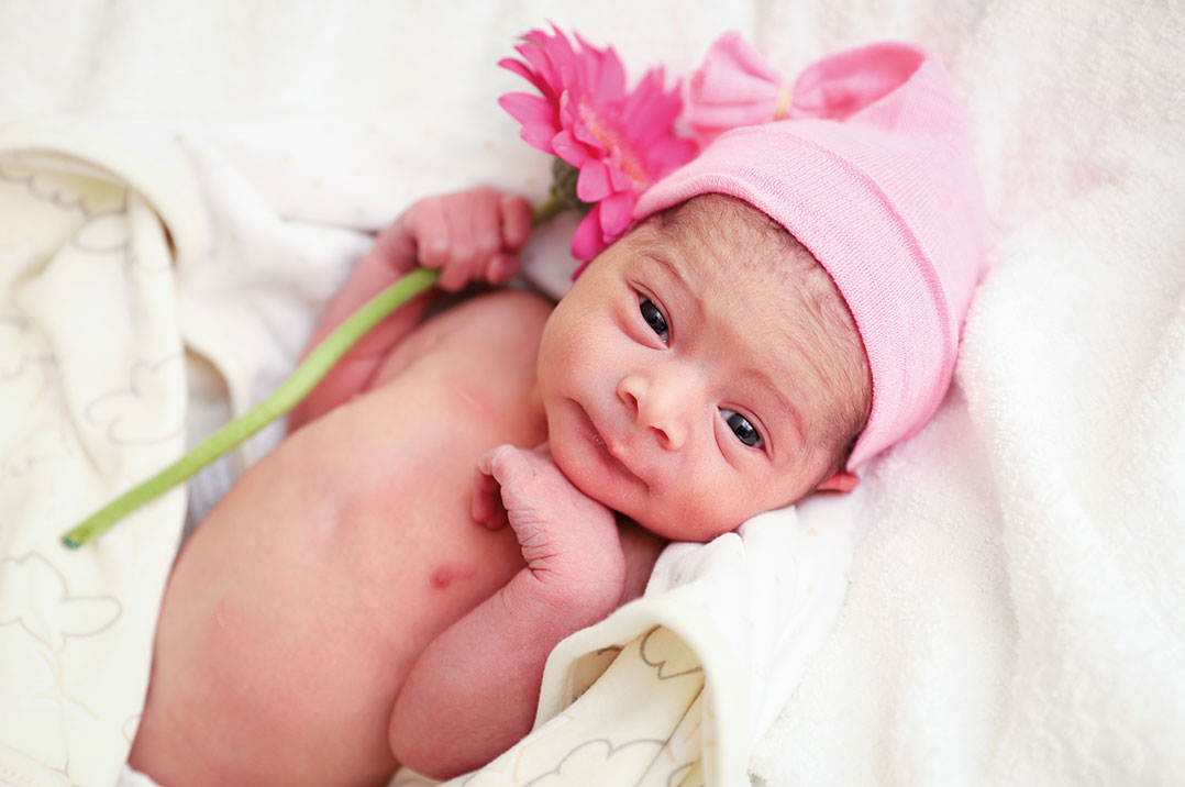 Newborn Esmie Jean DaCosta lying on a white blanket, wearing a pink cap and holding a pink flower