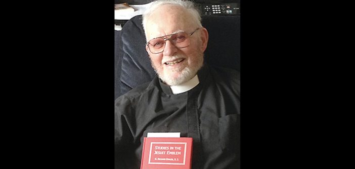 An elderly man wearing a black priest robe and holding a red book