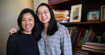 Two women stand and smile in front of a bookshelf.