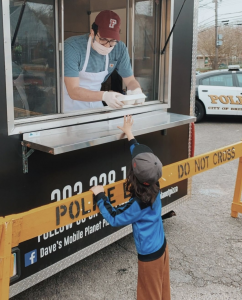 A young man wearing a maroon Fordham cap and standing in a food truck gives a tray of food to a little boy.