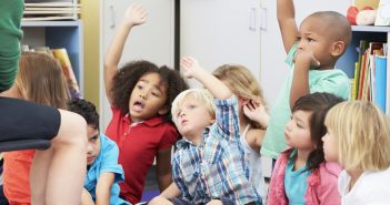 Children sit on a classroom carpet and excitedly raise their hands.