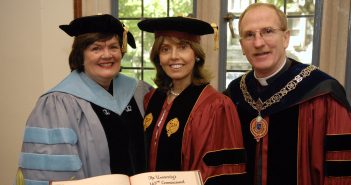 A woman wearing a blue graduation robe, a woman wearing a maroon graduation robe, and a man wearing a maroon graduation robe smile at the camera together.