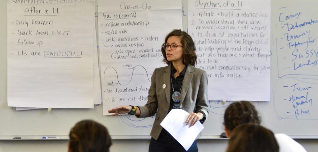A woman wearing glasses and a gray blazer stands in front of a dry erase board with writing on it.