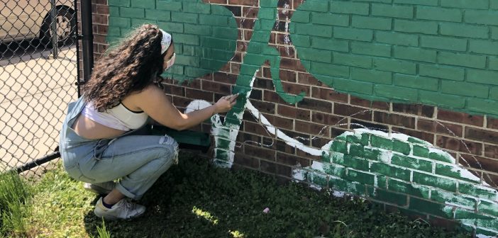 A girl wearing overalls and a mask squats in front of a brick mural and paints with a green brush.