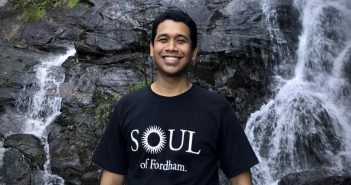 A man wearing a black t-shirt that says "Soul of Fordham" smiles in front of a waterfall.
