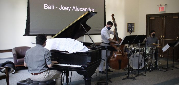 Three men each play an instrument: a piano, a cello, and a drum set, in a room with white walls.