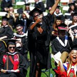 A woman wearing a black graduation gown stands, laughs, and holds up her phone to take a photo, while dozens of graduates around her sit and smile.