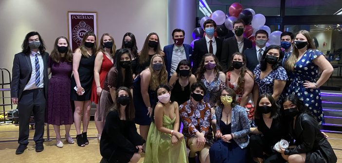 Twenty-four students wearing masks and formal attire smile for a group photo in an indoor setting.