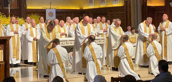 Four Jesuit priests laying their hands on four men who are being ordained priests, on the steps leading up tp the altar.