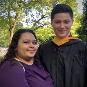 A woman wearing a purple shirt and a taller man wearing a black graduation gown smile.