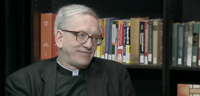 An elderly man wearing a black Jesuit shirt and glasses smiles in front of a bookshelf.