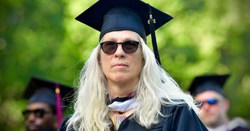 A woman with white blonde hair, sunglasses, and a graduation gown looks past the camera.