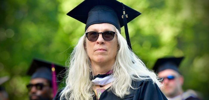 A woman with white blonde hair, sunglasses, and a graduation gown looks past the camera.