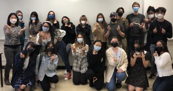 Twenty people wearing masks smile at the camera in front of a blank dry erase board and pose with their fingers in the shape of a heart.