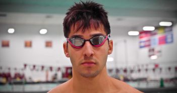 A man wearing goggles sternly looks at the camera in front of a swimming pool.