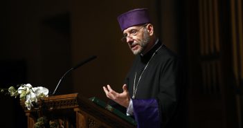 A man wearing a purple hat and black priestly robes holds his hand up at a podium.