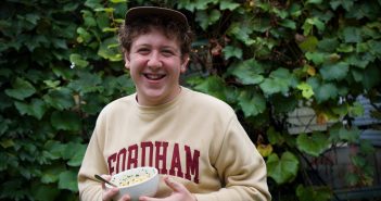 A boy holding a bowl of pasta smiles in front of an ivy-covered wall.