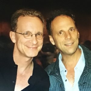 Two men smile next to each other in front of a dark background.