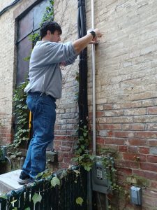 A man stands beside a brick wall and attaches a device to the side of the wall.