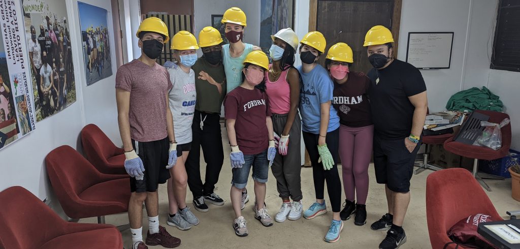 Nine students wearing masks and yellow construction hats smile in a building.