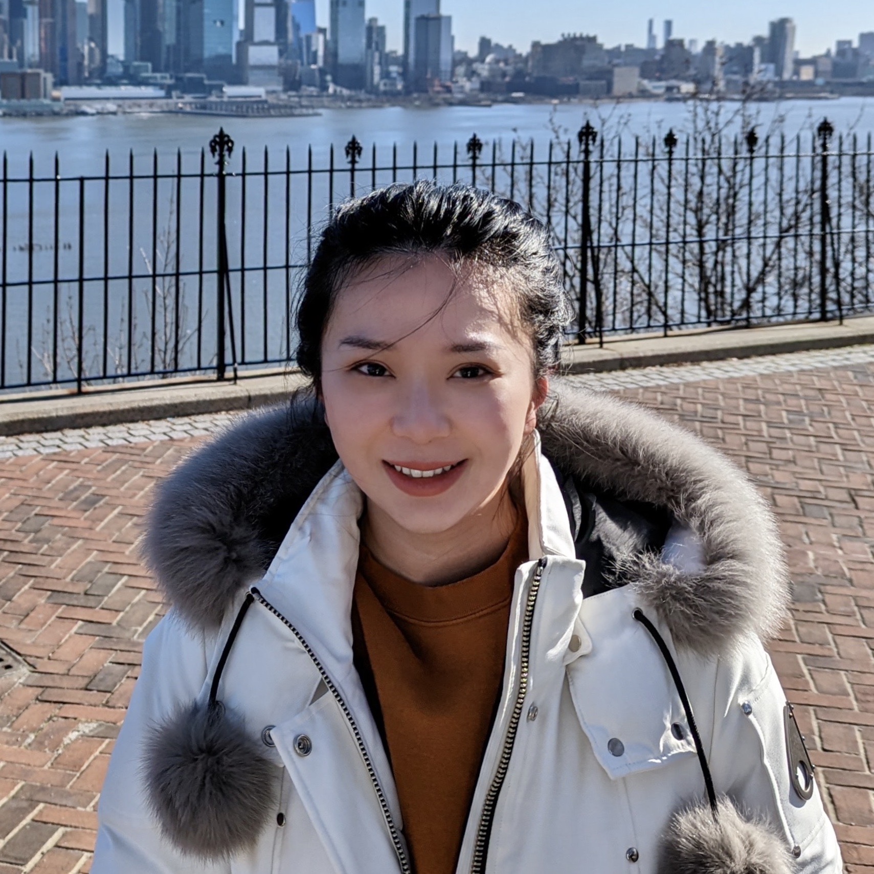 An Asian woman wearing a white hooded coat smiles in front of a city and a river skyline.