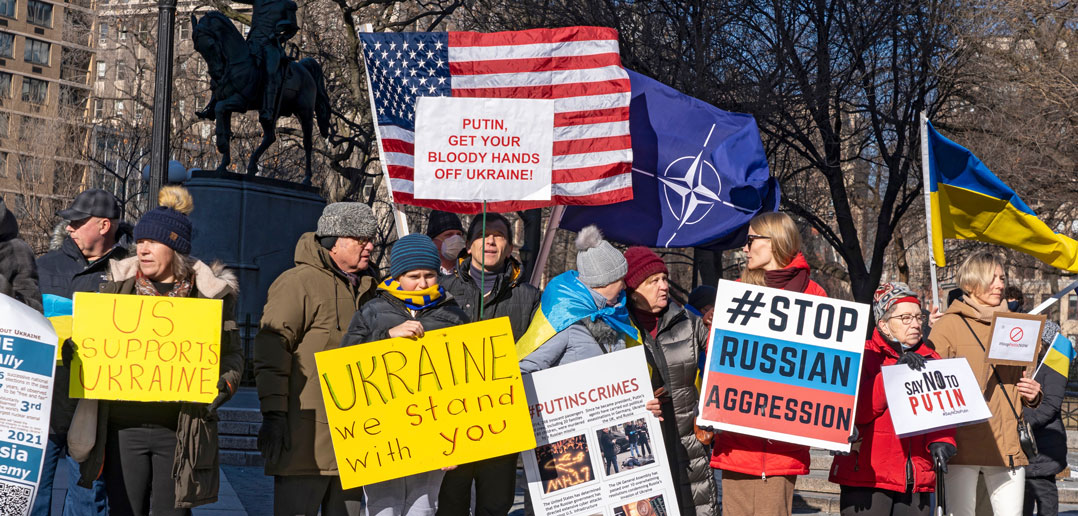 A group of people stand outside with colorful signs and an American flag, protesting against the Russian invasion of Ukraine.