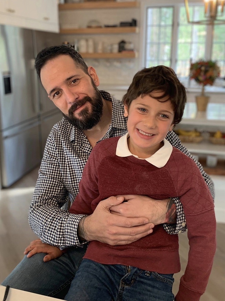A smiling man holds a smiling boy in his lap in a kitchen.