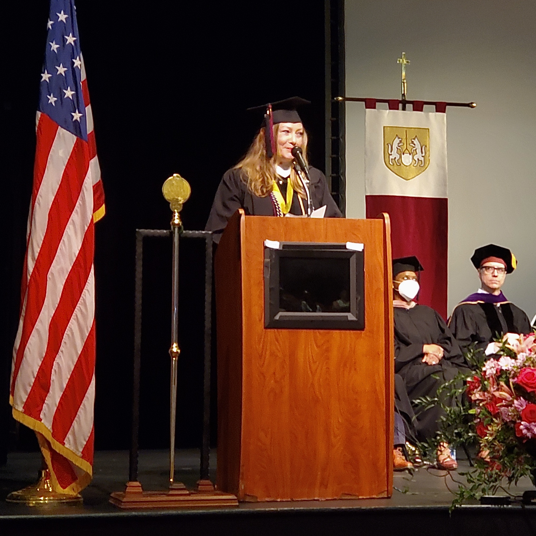 A woman wearing a graduation gown speaks in front of a podium.
