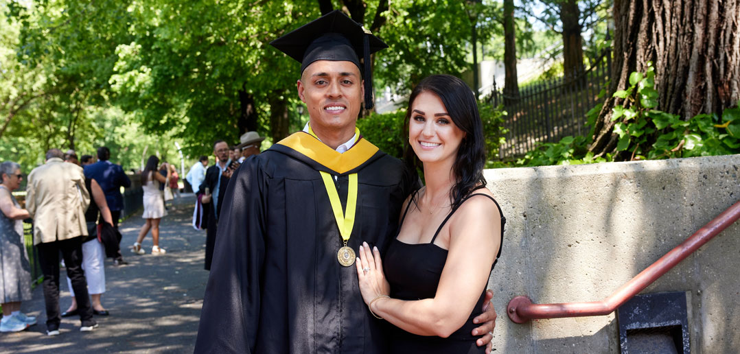 A man wearing a graduation gown smiles beside a woman wearing a black dress.