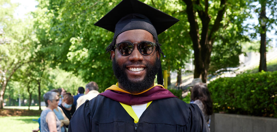 A man wearing sunglasses and a graduation gown smiles.
