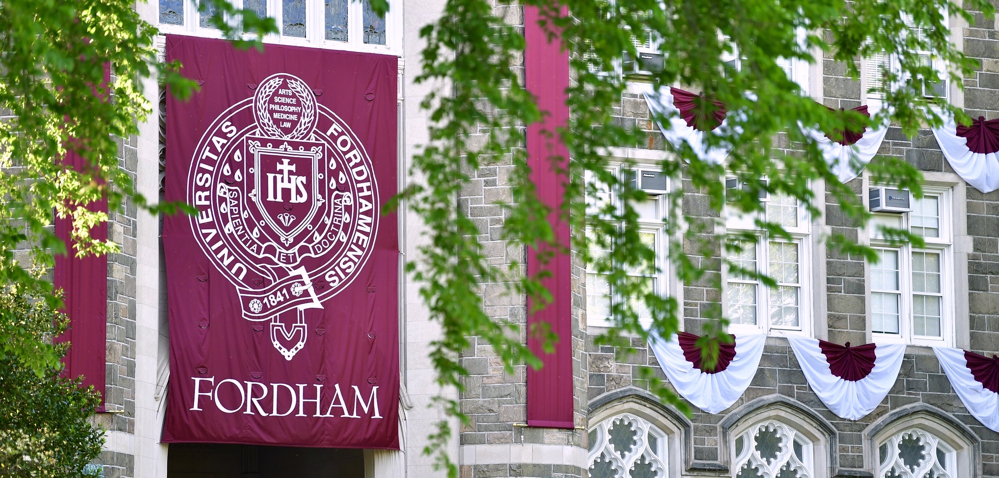 A maroon banner that says "Fordham" hangs in front of a building with leaves next to it.