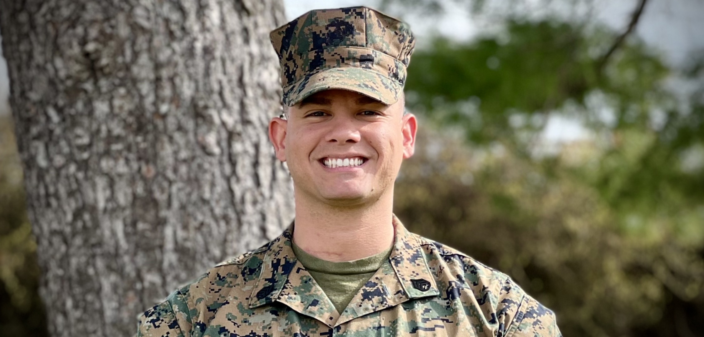 A man wearing a camouflage hat and shirt smiles in front of a tree.