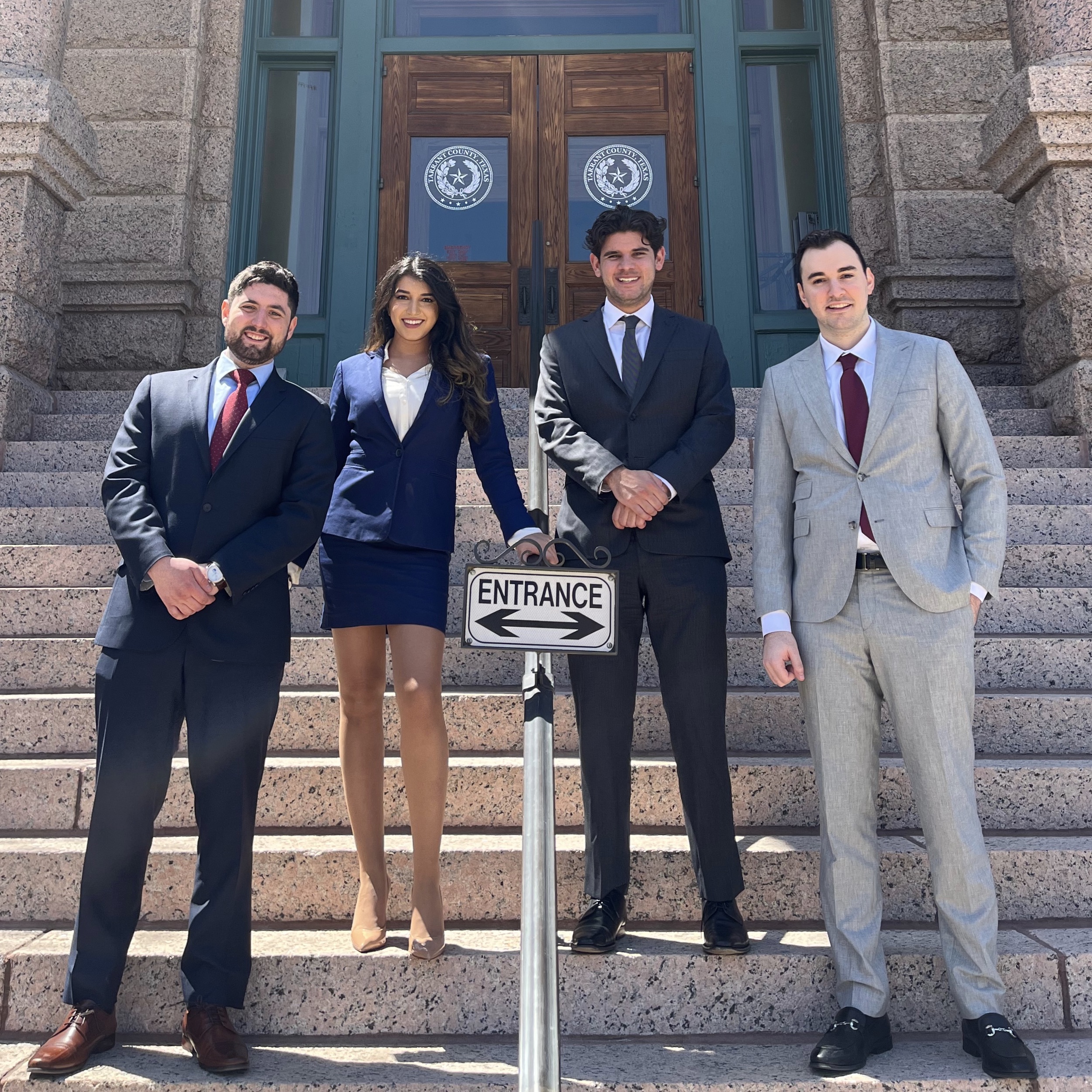 Four people wearing professional outfits smile on the steps of a courthouse.