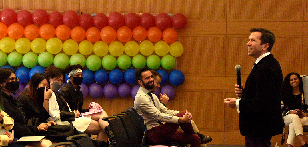 Patrick Hornbeck speaks to audience with rainbow colored balloons in bakcground
