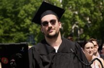 A man wearing sunglasses and a black graduation cap and gown smiles.